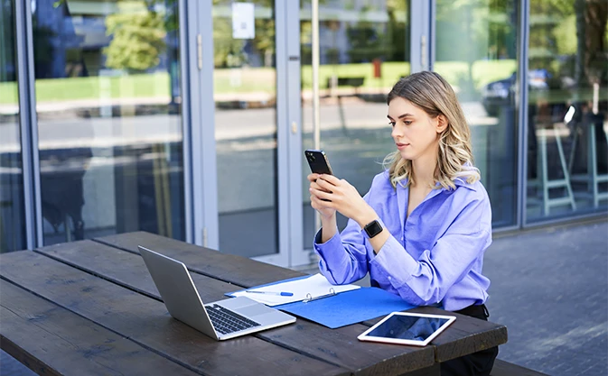 młoda kobieta pracująca zdalnie na świeżym powietrzu, patrząca na swój smartfon