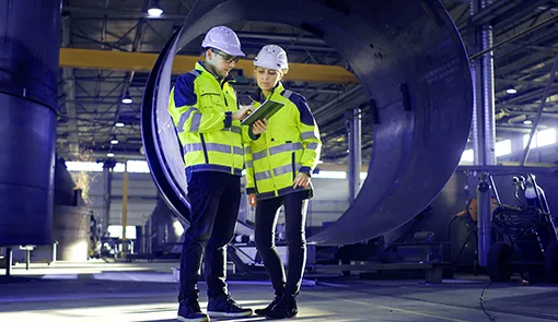 A mobility management project for manufacturing; a man and a woman are looking at a tablet screen, wearing work clothes and hard hats, with a production hall in the background